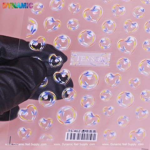 A hand in a black glove holds a sheet of heart-shaped, iridescent nail stickers featuring 3D bubbles. The sheet is labeled “3D Bubbles Iridescent (TS 462),” with product codes visible at the bottom. The pink background is adorned with Dynamic Nail Supply logos, enhancing its appeal.