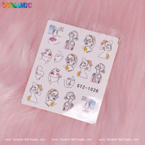 The sheet of decorative nail stickers showcases the Lady Face Hand writing art, featuring minimalist designs with colorful hats and accessories. Set against a backdrop of soft pink fuzzy fabric, the top corners display the word "Dynamic," and it is identified as STZ-1028 at the bottom, under the brand Dynamic Nail Supply.