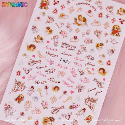 A white sheet titled "Angel face, Holy and Christian style (F427)" features a collection of decorative nail art stickers with Christian themes. The designs include cherubs, hearts, and roses, as well as scripted words like "love" and "forever." Set against a soft pink background is the Dynamic Nail Supply logo in the top left and bottom corners.