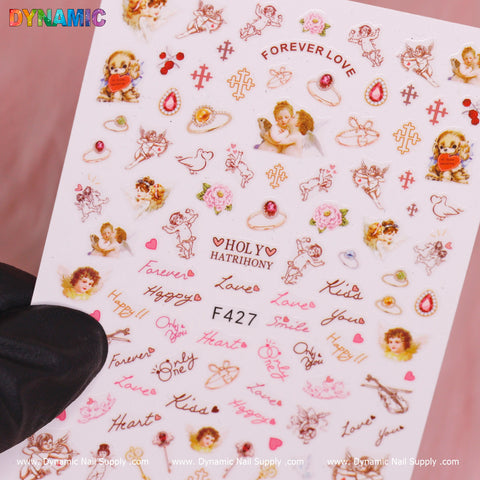A gloved hand holds a sheet labeled F427, featuring the Angel face, Holy and Christian style decorative nail stickers from Dynamic Nail Supply. The colorful stickers showcase cherubs, hearts, roses, swans, bows, and phrases like Forever Love, Kiss, Happy, and Holy Harmony. They come in various sizes with some adorned with rhinestones for added sparkle.