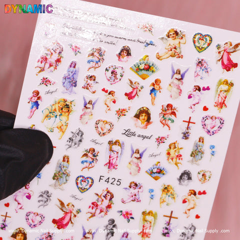 A sheet of vibrant Angel, Cupid, Holy and Christian style nail decals (F425) showcases cherubs, hearts, and flowers along with words like "Angel" and "Little Angel." A gloved hand presents the Cupid-inspired sheet against a pink background, with the Dynamic Nail Supply logo in rainbow colors at the top left corner.
