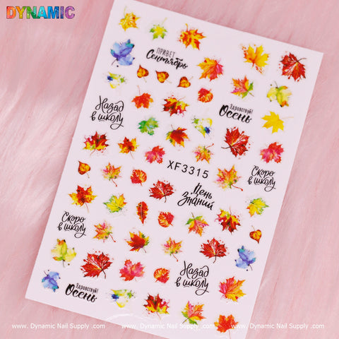 The "Fall Maple Leaves" sticker sheet, labeled XF 3315, showcases vibrant autumn-themed designs with colorful maple leaves and Russian phrases that include fall greetings and seasonal references. Set against a soft pink fabric background, the Dynamic Nail Supply brand name is elegantly displayed at the bottom edge.