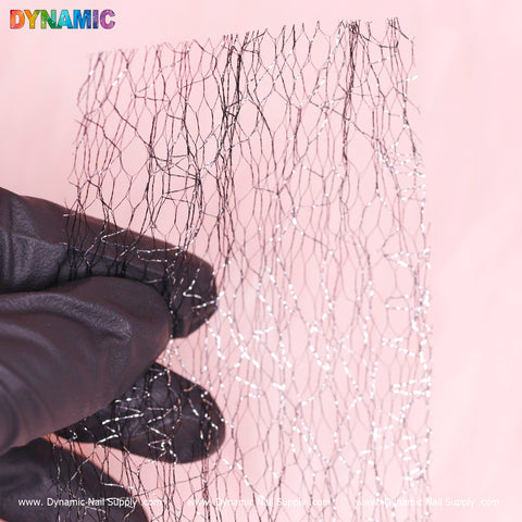 A hand in a black glove displays the Seamless Mesh Pattern—an abstract design featuring a complex mesh with shimmering silver strands—against a gentle pink backdrop. The word "DYNAMIC," styled in vibrant letters, adorns the top left corner, and website URLs are printed along the bottom edge. Presented by Dynamic Nail Supply.