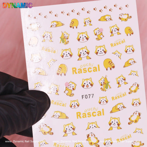 A sheet of nail art stickers from Dynamic Nail Supply's Ouchi Rascal Racoon Cartoon (F077) collection showcases cheerful yellow and white raccoon-like characters, some adorned with hats, surrounded by paw prints and the text "Ouchi Rascal." The sheet is partially held by a gloved hand against a pink background.