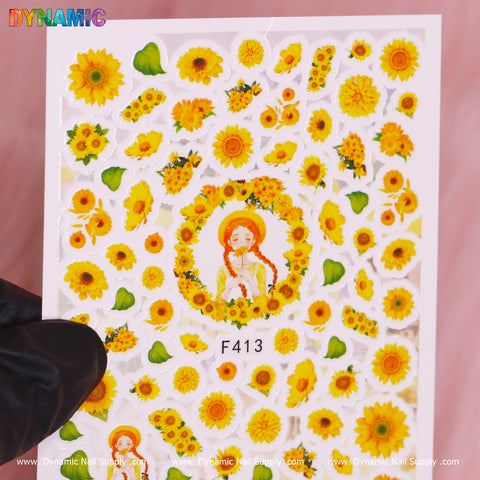 Held by a gloved hand against a pink backdrop, the "Sunflowers (F413)" sticker sheet from Dynamic Nail Supply bursts with vibrant sunflower designs, featuring individual blooms, clusters, and a figure encircled by sunflowers. The product showcases the dynamism of the sunflowers prominently marked with DYNAMIC at the top left corner.
