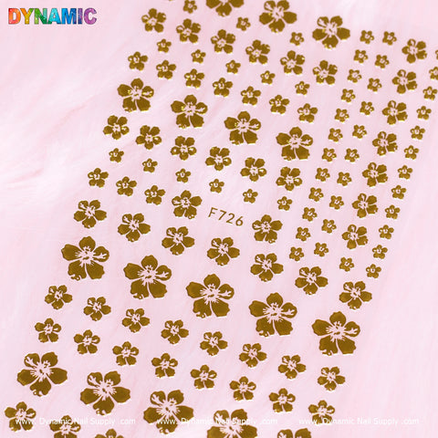 Gold Blossom Flowers Mixed-sizes (F726) nail art stickers embellish a soft pink backdrop, featuring gold blossom flowers of varying sizes that are arranged neatly in rows. The top of the image displays the brand name "Dynamic Nail Supply" in colorful letters.