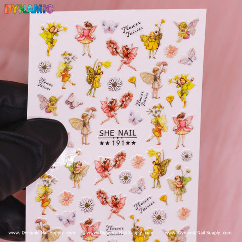 A hand in a black glove displays a sheet of whimsical nail stickers featuring spring fairies, butterflies, and daisies from Dynamic Nail Supply. These fairies showcase colorful wings and outfits in various poses. Set against a soft pink background, the Flower, Butterfly, Spring Fairies Sticker (191) is elegantly labeled in the center.
