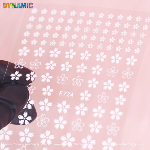 A black-gloved hand holds the "White Cherry Blossoms Mixed Sizes Sticker (F724)" from Dynamic Nail Supply, showcasing a grid of small white cherry blossom designs on a transparent sheet. The background features a soft pink hue, with "Dynamic Nail Supply" visible at both the bottom and top left corner.
