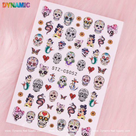 The Day of The Dead, Skull Sticker (STZ-CS052) by Dynamic Nail Supply is a vibrant sheet of nail art stickers adorned with sugar skulls, Day of the Dead motifs, pin-up mermaids, anchors, red roses, snakes, butterflies, and hearts with daggers. These striking designs stand out against a soft pink fur background and feature "DYNAMIC" in rainbow hues at the top left.