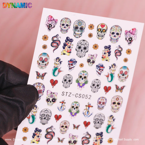 A gloved hand presents a dynamic sheet of nail art stickers from Dynamic Nail Supply, featuring Day of the Dead-themed designs like sugar skulls, whimsical characters, floral hearts, snakes, and anchors. This vibrant Skull Sticker set is identified by the code STZ-CS052 against a white backdrop.