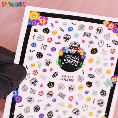 A hand in a black glove holds the Day of The Dead, Skull Sticker (CA-339) sheet from Dynamic Nail Supply, teeming with vibrant designs celebrating the Day of the Dead. Enjoy sugar skulls, flowers, ghosts, pumpkins, and phrases like Dia de los Muertos and Trick or Treat. The product code CA-339 COLOR is prominently displayed.