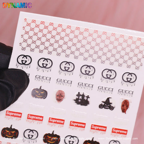 A hand in a black glove holds a Luxury Halloween Sticker (T-588) from Dynamic Nail Supply, featuring a variety of designs including repeated red GG patterns, black Gucci logos, jack-o-lanterns, skulls, Supreme logos, and other Halloween-themed images set against a light pink background.