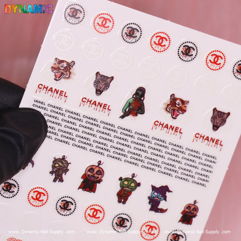 The Luxury Halloween Sticker (T-589) from Dynamic Nail Supply features an array of exquisite nail decals, such as Gothic-font "Chanel" lettering, images of wolves, stylized animals, and CC logos with a chic chain border. Ideal for creating a stylish Halloween appearance, the decals are set against a pink background and showcased by a gloved hand holding the sheet at its corner.