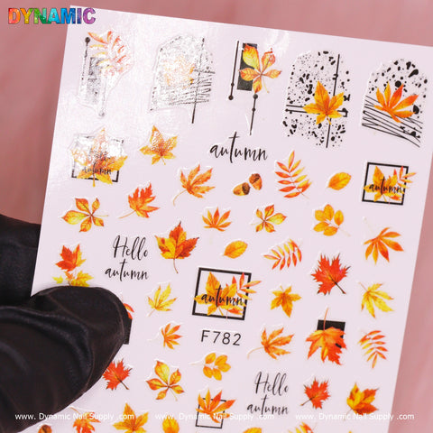 A gloved hand holds a sheet of Dynamic Nail Supply's Autumn, Fall Leaves Sticker (F782), showcasing vibrant fall leaves, acorns, branches, and abstract geometric patterns with "Hello Autumn" text in warm tones.