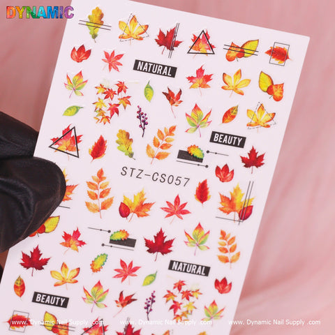 A sheet of Dynamic Nail Supply's Autumn, Fall Leaves Sticker (STZ-CS057) showcases vibrant nail art stickers adorned with red, orange, and yellow fall leaves. These autumn-inspired designs include geometric shapes and the words NATURAL and BEAUTY. A black-gloved hand presents the sheet against a soft pink backdrop.