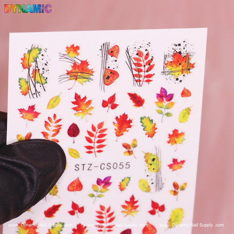 A hand in a black glove holds the Dynamic Nail Supply Autumn, Fall Leaves Sticker (STZ-CS055), which is filled with colorful fall leaves in shades of red, orange, yellow, and green, along with delicate butterflies, set against a soft pink background.