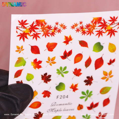 A black-gloved hand holds a sheet of vibrant Autumn, Fall Leaves Sticker (F204) featuring red maple leaves along with green, yellow, and orange leaves. Branded by Dynamic Nail Supply, this product captures the essence of autumn.