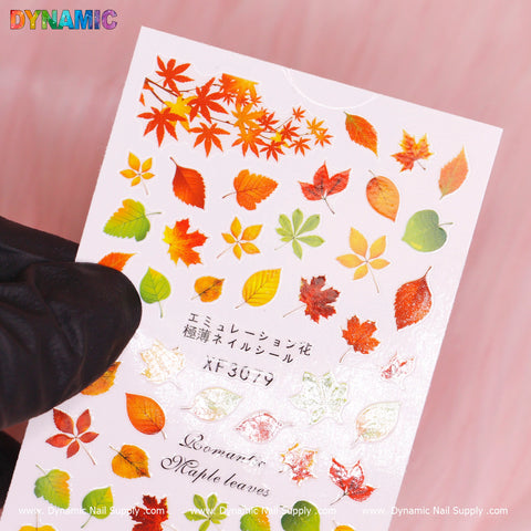 A close-up of the Autumn, Fall Leaves Sticker (XF3079) by Dynamic Nail Supply displays a variety of colorful autumn leaves, featuring maple and oak designs in red, orange, yellow, and green hues. A gloved hand holds the fall-themed sticker sheet against a pink background. Text in English and Japanese is visible alongside the product code XF3079.