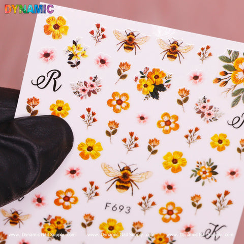 A hand wearing a black glove holds a sheet labeled F693, featuring Spring Time Flower Stickers from Dynamic Nail Supply. The stickers showcase intricate bees, vibrant orange and yellow flowers, and decorative letters set against a soft pink background.