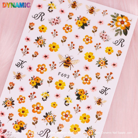 A sheet of vibrant Spring Time Flower Stickers from Dynamic Nail Supply, featuring floral designs, buzzing bees, and the decorative letters K and R. The flowers bloom in shades of orange, yellow, and pink against a soft pink background. Labeled F693, the brand's name stands proudly at the top.