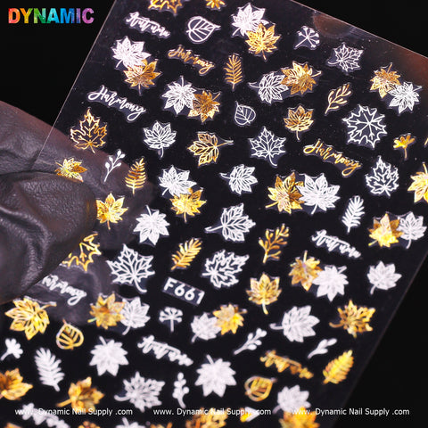 A gloved hand is displaying a sheet of Gold And White Autumn Leaves Stickers (F661), featuring an elegant design with gold and white maple leaves, the word "Harmony," and acorns. The branding of Dynamic Nail Supply appears at both the top and bottom.