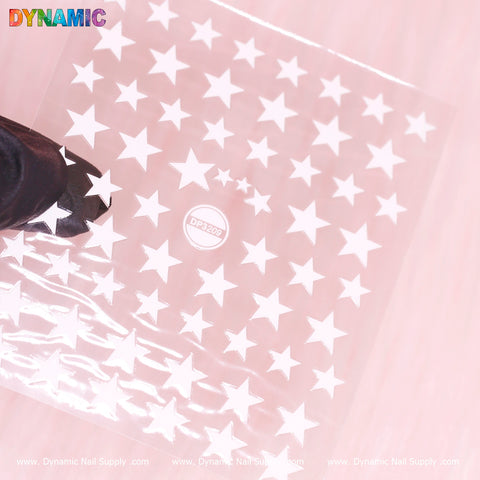 Clear plastic nail stencil showcasing an array of white star stickers and cutouts, positioned on a pink surface and gently held by a hand. The vibrant DYNAMIC logo is displayed at the top left corner, with DynamicNailSupply.com repeated along the bottom edge. Product Model: White Star Sticker (DP3209) by Dynamic Nail Supply.