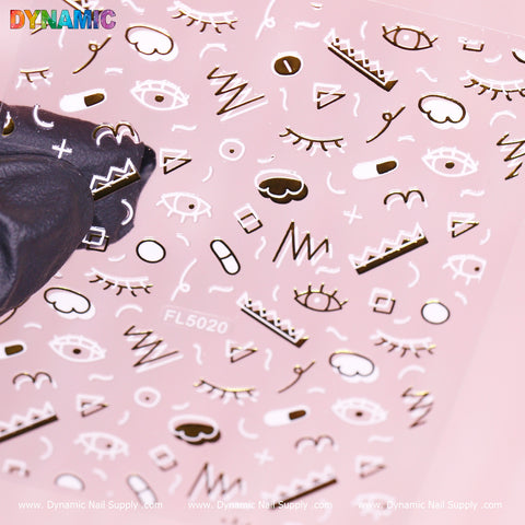 A sheet of Hand-drawing Sticker (FL5020) by Dynamic Nail Supply showcases abstract designs with eyes, lips, geometric shapes, and wavy lines in white and metallic gold on a pink background. A black gloved hand holds the sheet from the left side, displaying the product code FL5020 at the bottom.