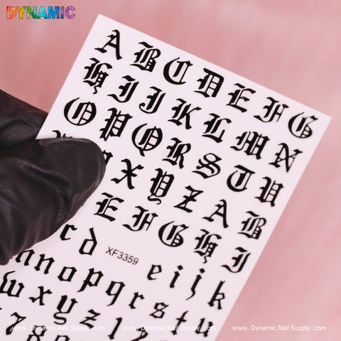 A gloved hand holds a sheet of Roman Letter and Number Stickers (XF3359) from Dynamic Nail Supply, featuring black Gothic letters and numbers on a pink background. The set includes both uppercase and lowercase stickers, making them perfect for any stylish manicure.