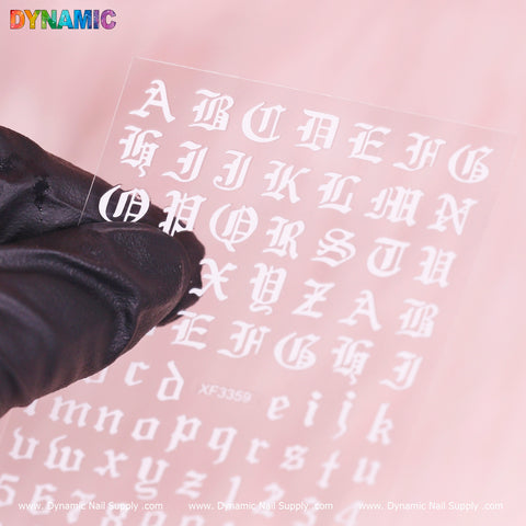 A gloved hand holds a sheet of the Roman Letter and Number Sticker (XF3359) against a pink background. The stickers feature Gothic-style uppercase and lowercase letters in white, with the words "DYNAMIC" and "Dynamic Nail Supply" printed at the top and bottom edges of the image.