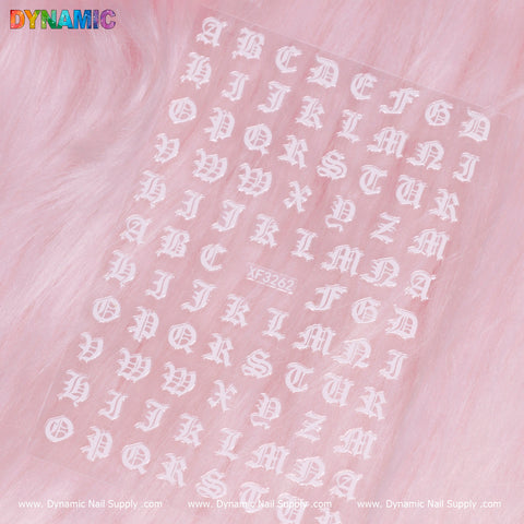 On a textured pink background, a sheet of white Roman Letter Sticker (XF3262) from Dynamic Nail Supply is displayed. The Roman-style alphabet stickers are neatly arranged in rows, with the brand name "Dynamic" prominently featured in the top left corner.