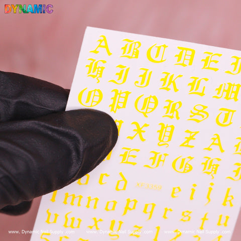 A gloved hand holds a white sheet displaying uppercase and lowercase stickers of yellow Gothic-style Roman letters from the "Roman Letter and Number Sticker (XF3359) - Yellow" collection by Dynamic Nail Supply, set against a pink background. The top left corner showcases the brand name DYNAMIC in vibrant text.