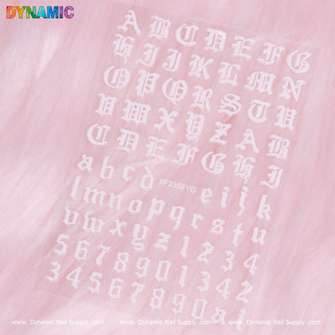 A sheet of the Letter and Number Sticker (XF3359YG) from Dynamic Nail Supply is displayed on a pink fluffy background. The stickers showcase uppercase and lowercase letters, as well as numbers 0-9, in a stylized ornate font. The Dynamic Nail Supply logo appears at both the top and bottom of the image.