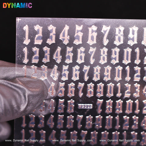 A gloved hand displays a transparent sheet featuring silver, Gothic-style numbers aligned in rows, accentuated by the reflective nature of the Number Sticker (WG623) - rose on a rose-hued background. "DYNAMIC" is prominently displayed at the top in vibrant letters, with "Dynamic Nail Supply" repeated along the bottom edge.