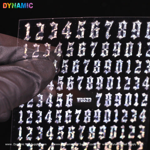 A hand wearing a black glove holds a sheet of silver, metallic number stickers with a gothic font, featuring numbers from 0 to 9 arranged neatly in rows. The shiny texture of these Number Stickers (WG623) - Silver is highlighted against the dark background. The brand name, Dynamic Nail Supply, prominently appears at the top left, adding an aura of mystery.