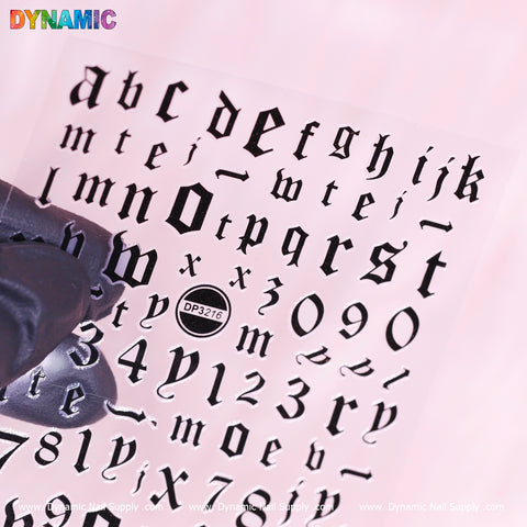 A gloved hand holds a transparent sheet displaying the Letter and Number Sticker (DP3216) - Black from Dynamic Nail Supply, featuring Gothic-style letters a to z and numbers 0 to 9. The top left corner spells out "DYNAMIC" in multicolored letters, while the bottom edge is repeatedly branded with "Dynamic Nail Supply," and DP3216 is subtly marked on the side.