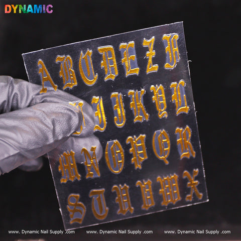 A gloved hand holds a transparent sheet of Roman Alphabetical Sticker (Gold) in Gothic style. The dark background accentuates their metallic sheen. "Dynamic" is printed in multicolored letters at the top left, with www.DynamicNailSupply.com along the bottom edge from Dynamic Nail Supply.