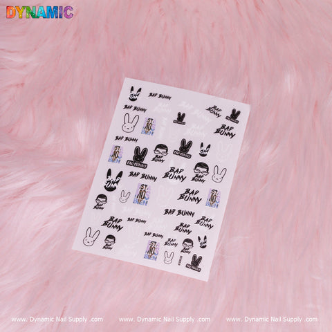 A sticker sheet featuring a variety of black and white designs, including bunny silhouettes and the Hip Hop Sticker style 'Bad Bunny' text from Dynamic Nail Supply. The colorful graphics are set against a pink fluffy backdrop, with the Dynamic Nail Supply brand name repeatedly printed along the bottom edge.