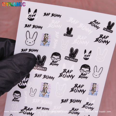 A person wearing a black glove holds a sheet of Dynamic Nail Supply's "Bad Bunny, Hip Hop Sticker (MG10825-30) - Black and White" nail art decals. The sheet features hip hop stickers like Bad Bunny text, a stylized bunny face, and an image of a person in sunglasses. With the phrase "Está No Bien" shining brightly, the sheet stands out against a blurred pink background.