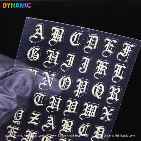 A gloved hand holds a transparent sheet showcasing the Roman Alphabetical Sticker (Silver) from Dynamic Nail Supply. Each Gothic font letter, adorned with decorative flourishes on a clear background, is perfect for nail art application. The brand name, Dynamic Nail Supply, is repeated along the bottom edge of the sheet.