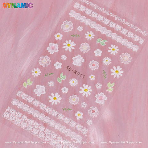 A pink background showcases a transparent sheet of nail decals, labeled as Floral Sticker (5D-K011). It is adorned with white lace patterns and vibrant floral designs in shades of white, pink, yellow, and green. The borders are elegantly framed by the multicolored text logos of Dynamic Nail Supply.