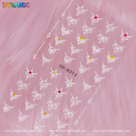A sheet of Design Nail Sticker (5D-K013) rests on a pink background, displaying delicate white lace-like patterns embellished with vibrant rhinestones in shades of yellow, pink, and orange. The 5D-K013 design code is prominently featured alongside the Dynamic Nail Supply logo at the top.