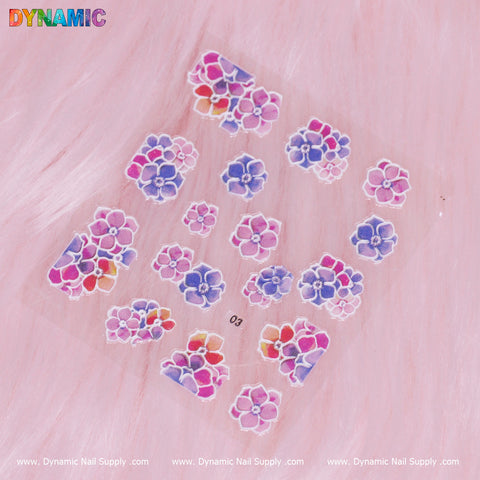 Against a pink, fluffy background, a sheet of vibrant stickers showcases colorful flower designs. These blossoms come in shades of purple, pink, blue, and orange and are arranged in clusters. Below them is the text "Dynamic Nail Supply" with "Flower Sticker (03)" printed near the center.