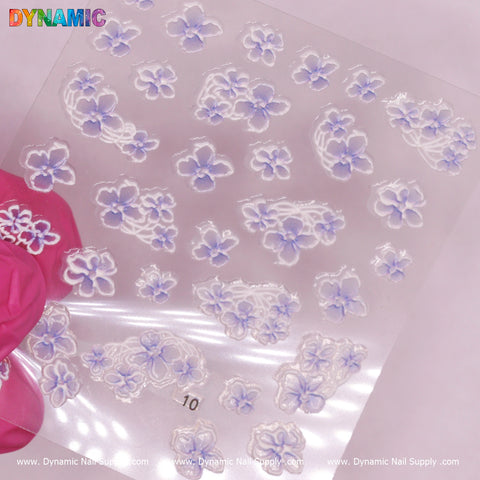 A pink-gloved hand displays a transparent sheet of Flower Sticker (10) from Dynamic Nail Supply, featuring delicate white flowers with light purple centers and various petal arrangements. The background has a subtle grid design, and the brand name is visible at the bottom, emphasizing this exquisite sticker pack.