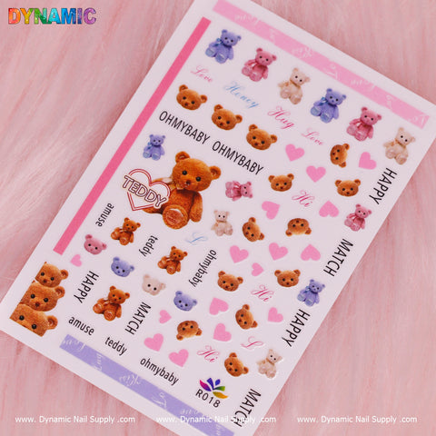 A sheet of the Cute Teddy Bear Stickers (R018) from Dynamic Nail Supply rests on a pink furry backdrop. Cartoon teddy bears in brown, purple, and pink are featured alongside words like Teddy, OHMYBABY, HAPPY, and LOVE in shades of pink and purple. The brand Dynamic Nail Supply is clearly displayed on this adorable sticker set.