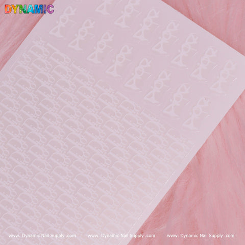 A luxurious sheet of Dynamic Nail Supply's "Luxury Water Decal (White)" nail art stickers, featuring intricate designs and bow-shaped details, is displayed on a soft pink, fur-like surface. This elegant collection is accented by the text "DYNAMIC" along with the website www.DynamicNailSupply.com on its edge.