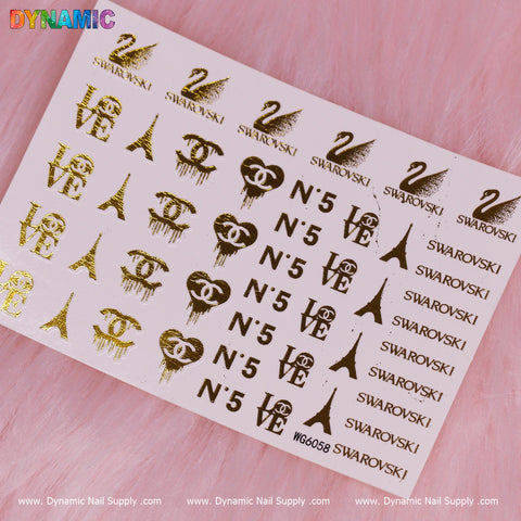 A luxurious sheet of metallic gold decals from the "Luxury Style Water Decal (WG6058) - Gold" by Dynamic Nail Supply lies on a pink furry backdrop. The design features symbols such as the Eiffel Tower, LOVE, N°5, SWAROVSKI, and a stylized double C. In the top left corner, "DYNAMIC" appears in rainbow letters, while the bottom displays Dynamic Nail Supply's website.