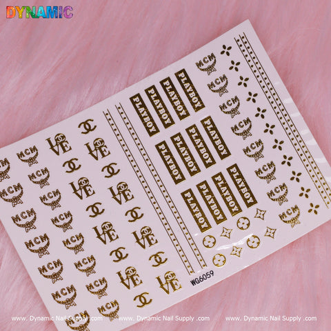 A luxurious sheet of gold water decals from Dynamic Nail Supply's Luxury Style Water Decal collection (WG6059) is set against a pink textured background. It features logos and symbols such as MCM, Playboy, LOVE, interlocking CC motifs, and geometric shapes.