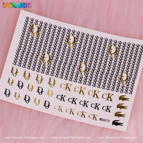 A white sheet of Luxury Style Water Decal (WG6073) from Dynamic Nail Supply is displayed against a pink background, highlighting various brand-inspired nail designs. It showcases black and gold interlocking letters resembling famous fashion logos, featuring patterns such as D, CK, and small crocodile images.