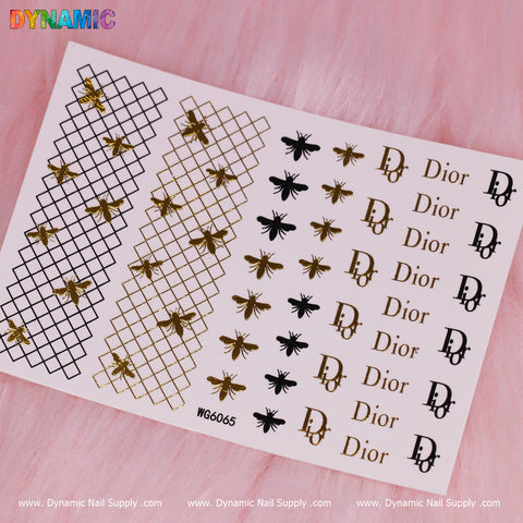 The Luxury Style Water Decal (WG6065) sheet from Dynamic Nail Supply, adorned with gold and black bees, Dior logos, and crisscross patterns, lies on a pink textured background.