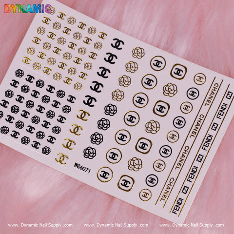 A sheet of Luxury Style Water Decal (WG6071) from Dynamic Nail Supply is elegantly displayed on a pink, furry surface. Featuring black and gold logos such as interlocking C designs, flowers, and the words Fendi and Chanel, it encapsulates sophistication.